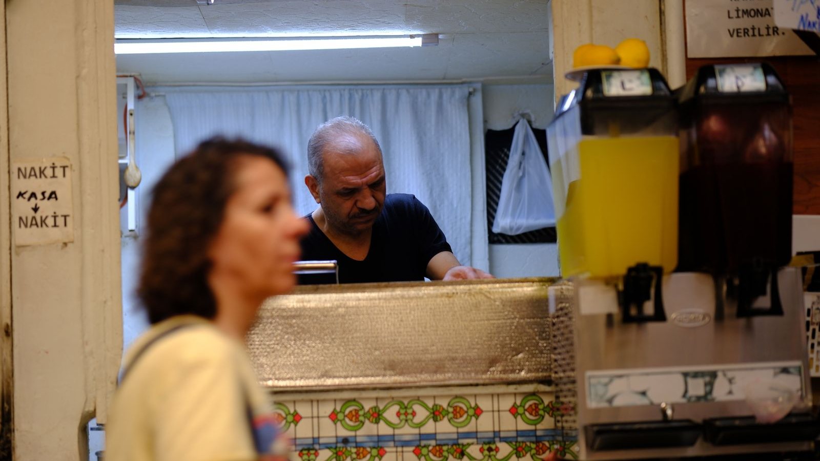 Küçük bir esnaf dükkânının tezgâhı; duvarda “nakit kasa” yazan el yazısı bir levha.