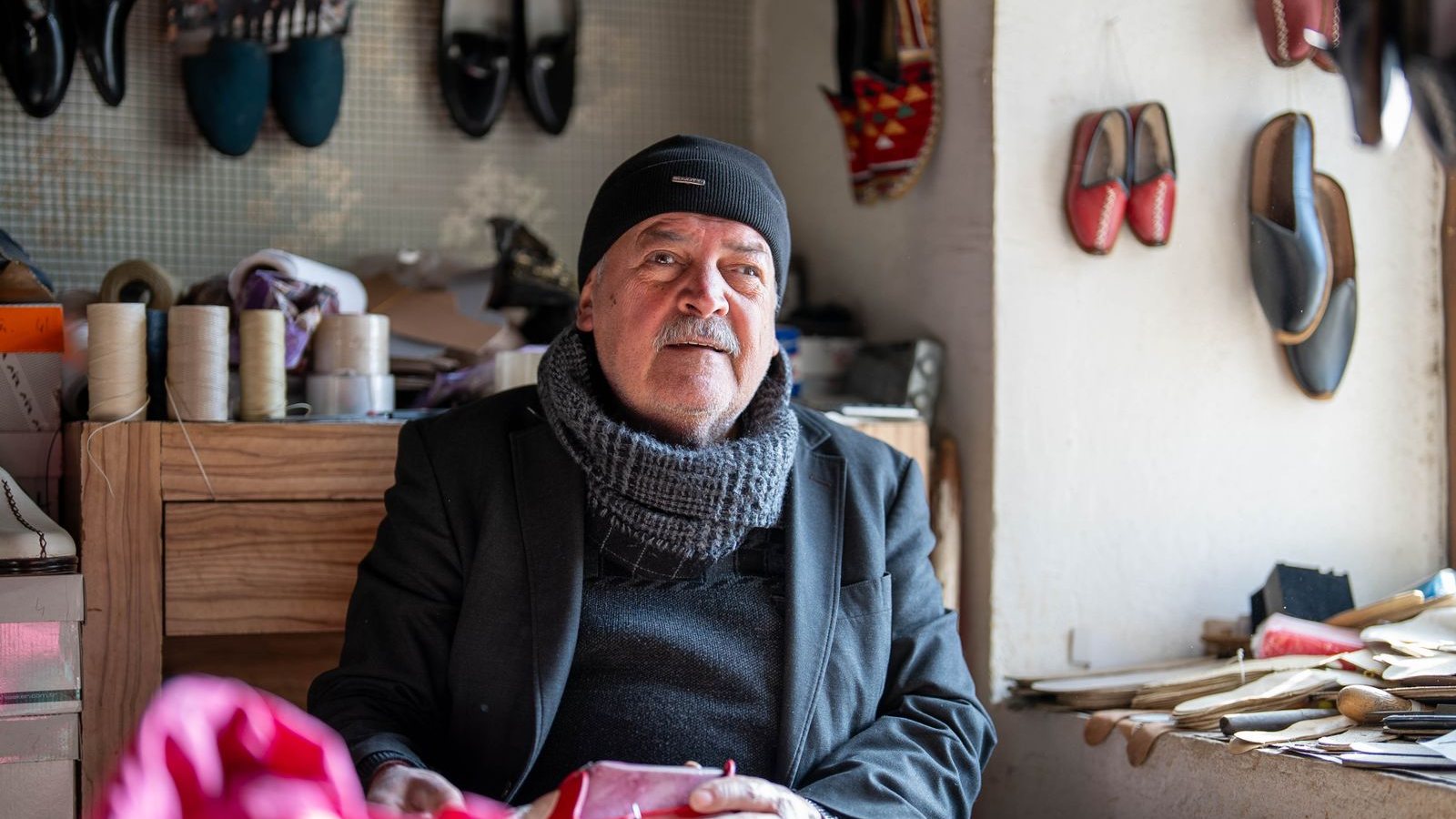 Atölyesinde elinde işlediği ayakkabıyla oturan yaşlı ayakkabı ustası; duvarda ürettiği modeller asılı.